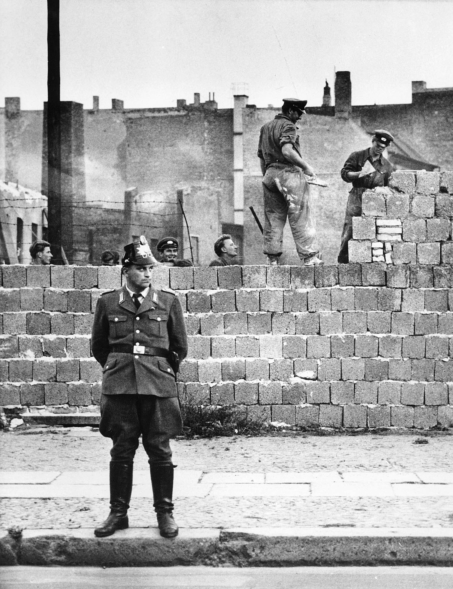 Policajt zo Západného Berlína stojí pred betónovým múrom oddeľujúcim východný a západný Berlín na Bernauer Strasse, zatiaľ čo robotníci z východného Berlína pracujú na stavbe múru