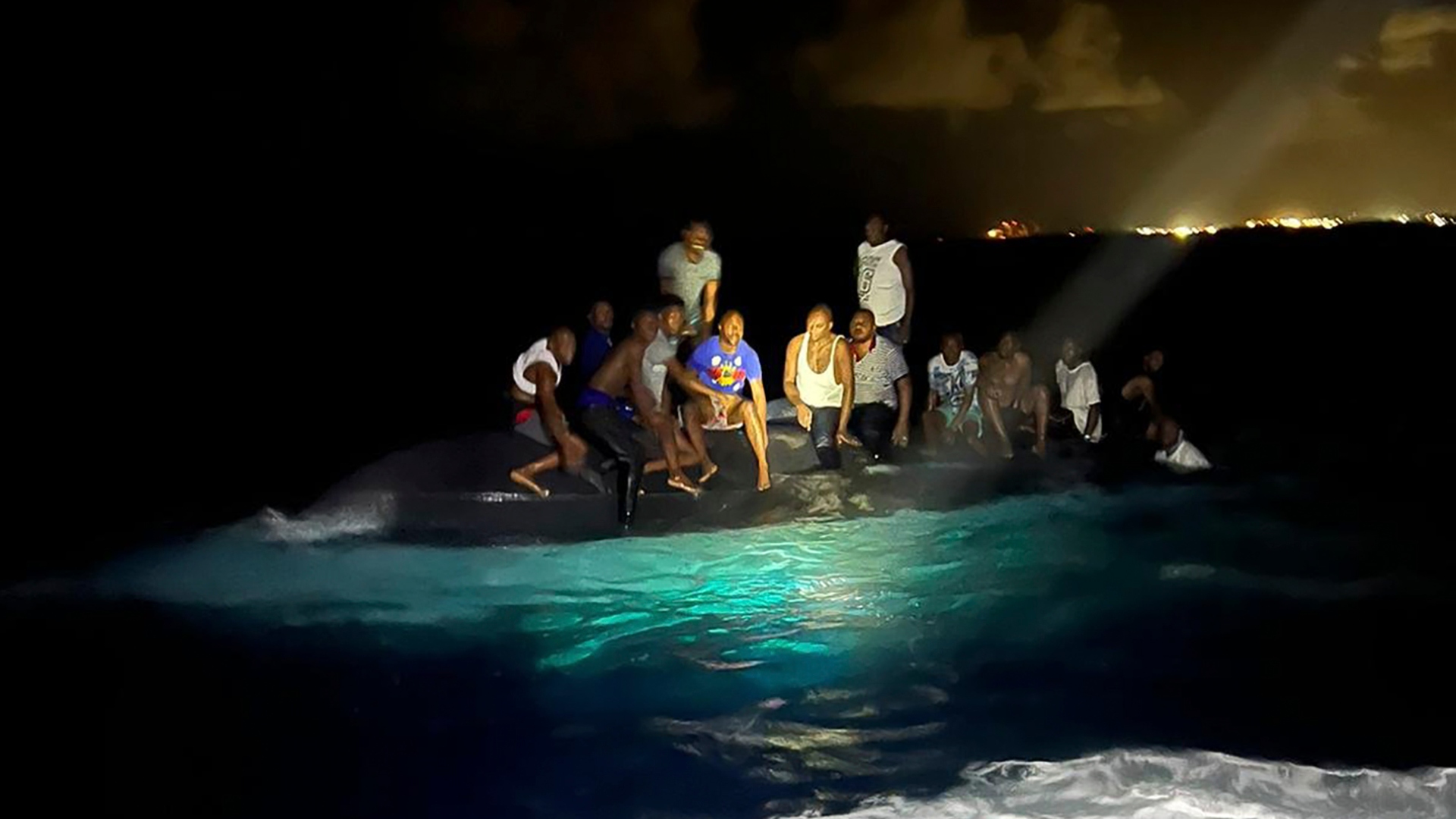 Na snímke preživší migranti z Haiti sedia na prevrátenom člne  neďaleko New Providence na Bahamách v nedeľu 24. júla 2022. Bezpečnostné zložky našli 17 mŕtvych tiel po tom, ako sa v nedeľu ráno pri pobreží Bahám v oblasti Karibského mora prevrátilo plavidlo s migrantmi z Haiti.