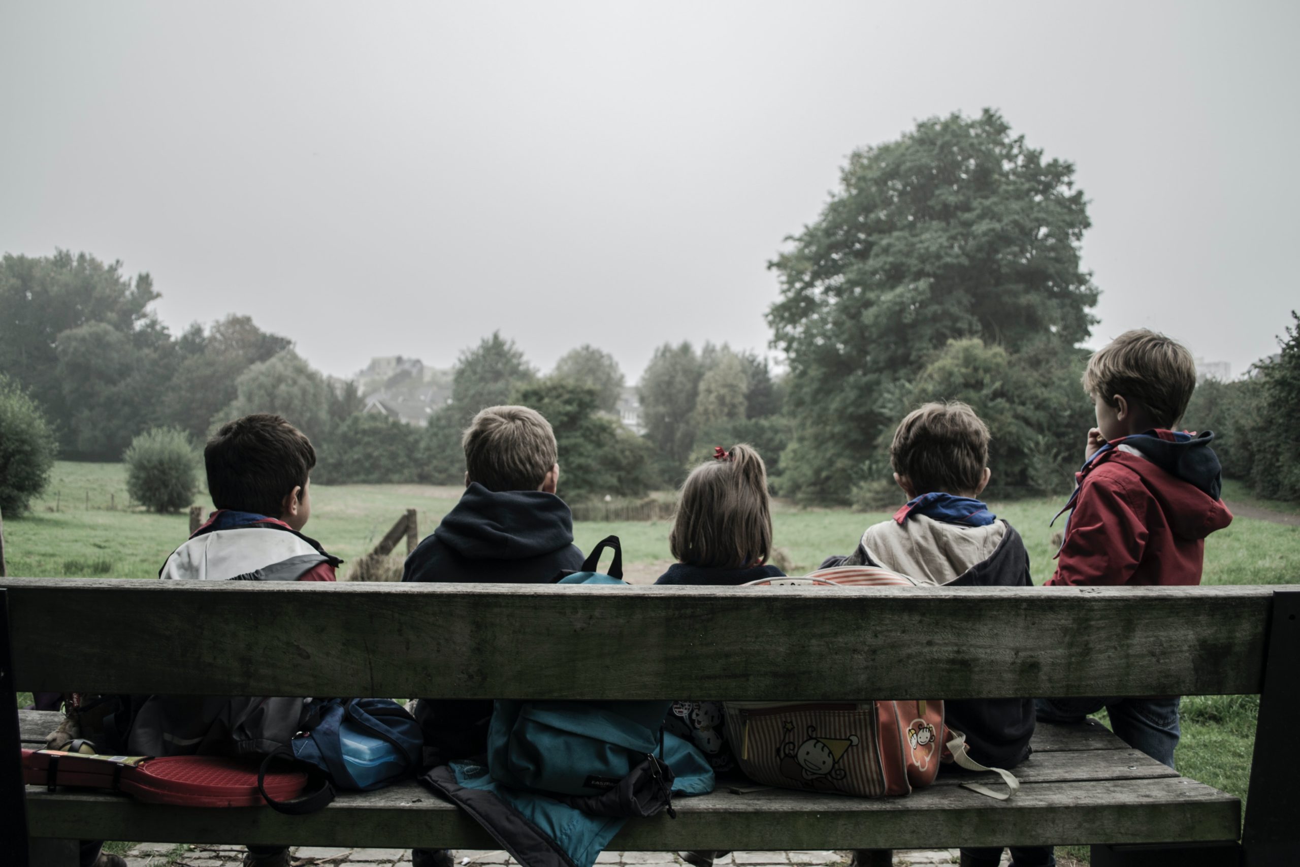 5 detí so školskými taškami sedia na lavičke otočené chrbtom a pozerajú na lúku a stromy