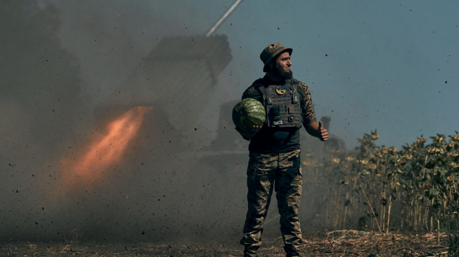 Ukrajinský vojak drží melón v Donecku, zatiaľ čo v pozadí strieľa salvový raketomet (MLRS).
