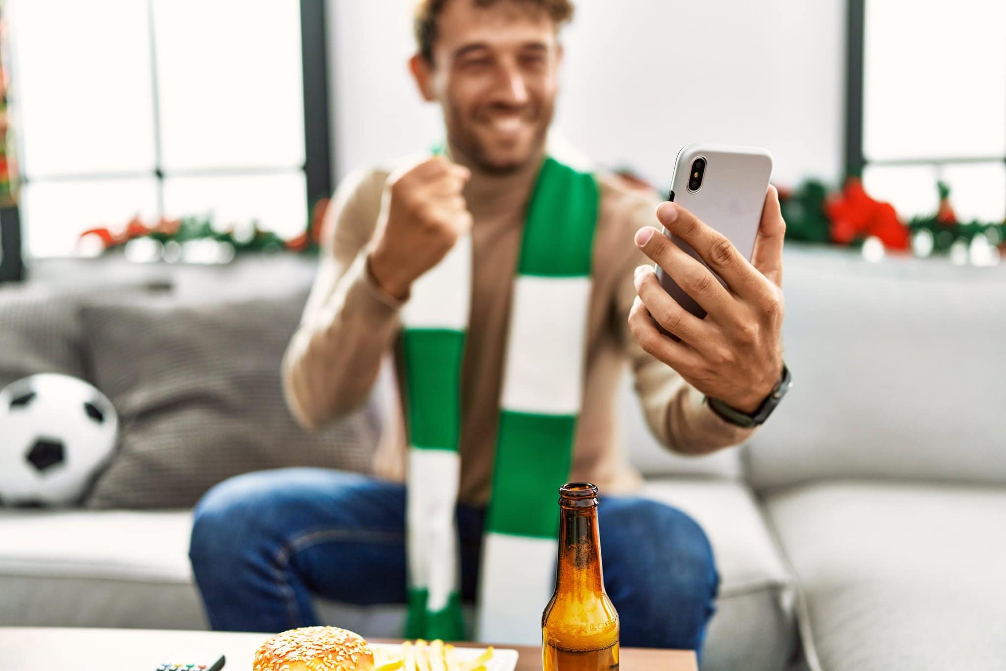 young-hispanic-man-supporting-soccer-match-using-smartphone-sitting-by-christmas-decor-home