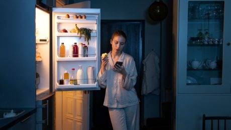 Žena stojí pri otvorenej chladničke a pozerá do mobilu.